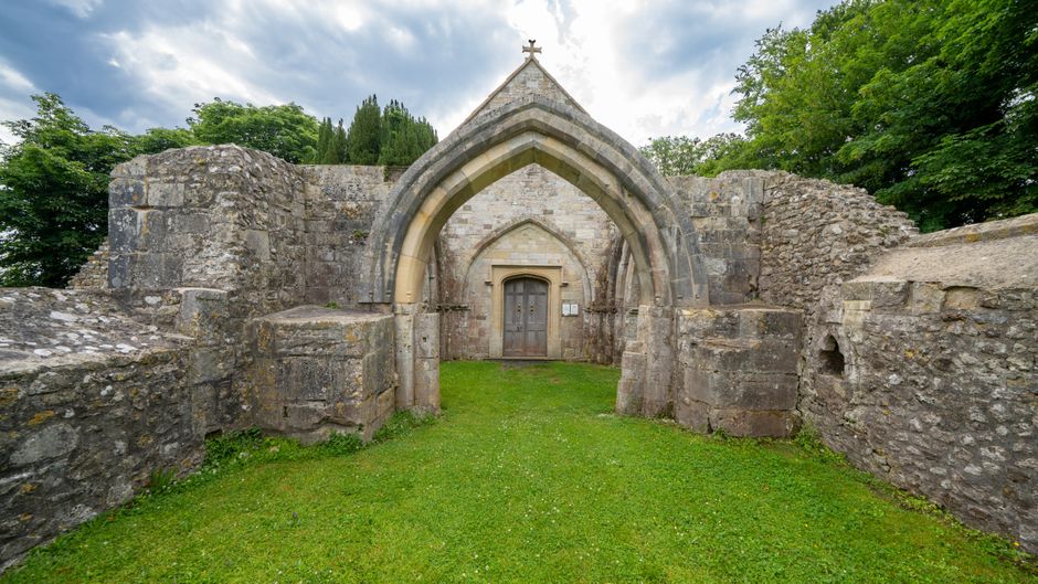 The image shows the entrance to an ancient stone church or chapel, featuring a pointed Gothic archway with a wooden door set into the stone facade. The structure is surrounded by low stone walls and appears to be in a grassy area, with lush green trees and foliage in the background. The Gothic arch is constructed from large, weathered stone blocks, with a small cross mounted at the top of the arch. To the right, part of the stone wall extends outward, and on the left, the wall curves inward. The scene is well-lit with natural daylight, highlighting the textures of the stonework and the vibrant green grass below. The setting suggests a historic site, possibly a medieval church or ruin, with a peaceful and well-maintained environment. Sutton Man and Van would assist with house removals or furniture transport if relocating near historic landmarks like this, supporting their connection to local heritage and logistics services.