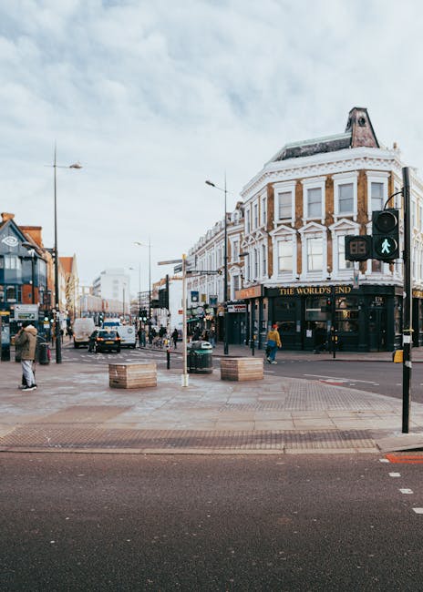 A street view on Sutton High Street showing a busy pedestrian area with a traffic light displaying a green pedestrian signal, several people walking across the crosswalk, and others standing or walking along the sidewalk. The scene includes parked cars along the curb, a double-decker bus on the left, and a variety of shopfronts including The World's End pub on a classic white building with bay windows. The foreground features a paved pedestrian crossing with large wooden pallets placed on the sidewalk, indicating preparations for home relocation or furniture transport. The lighting suggests daytime with a partly cloudy sky, and the overall environment reflects an urban setting suitable for moving or packing services offered by Sutton Man and Van.